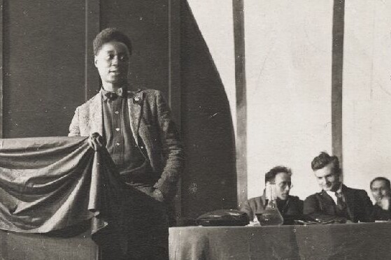 Claude McKay standing at a lectern with people seated at a long table behind him
