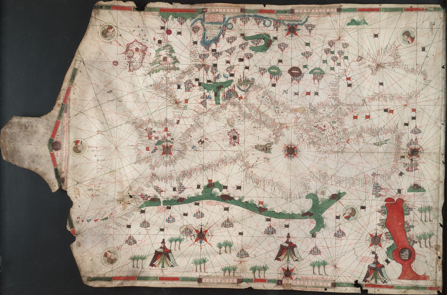 A portolan chart. A large map of lands surrounding the Mediterranean Sea on a single piece of parchment. There are green mountains and the Red Sea is colored red. There are faces blowing winds from sixteen directions toward the center of the map.