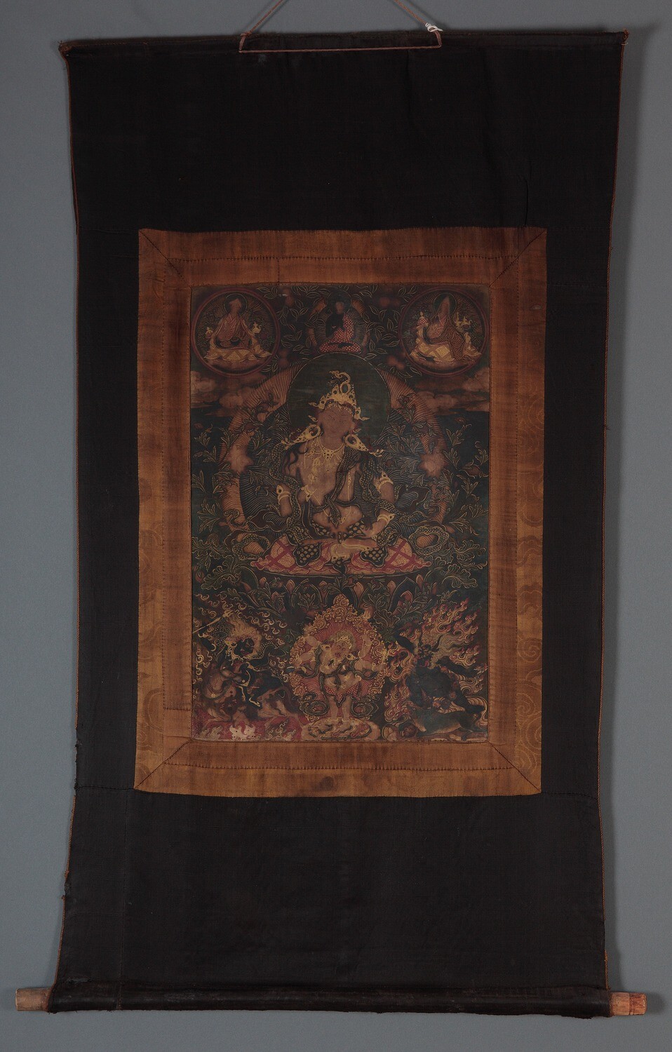 Tibet MSS 62, Box 72. Tanka painting mounted on dark colored fabric support. A figure is seated in the center of the painting, adorned with gold details. Six smaller figures, three above and three below, surround the central subject. 